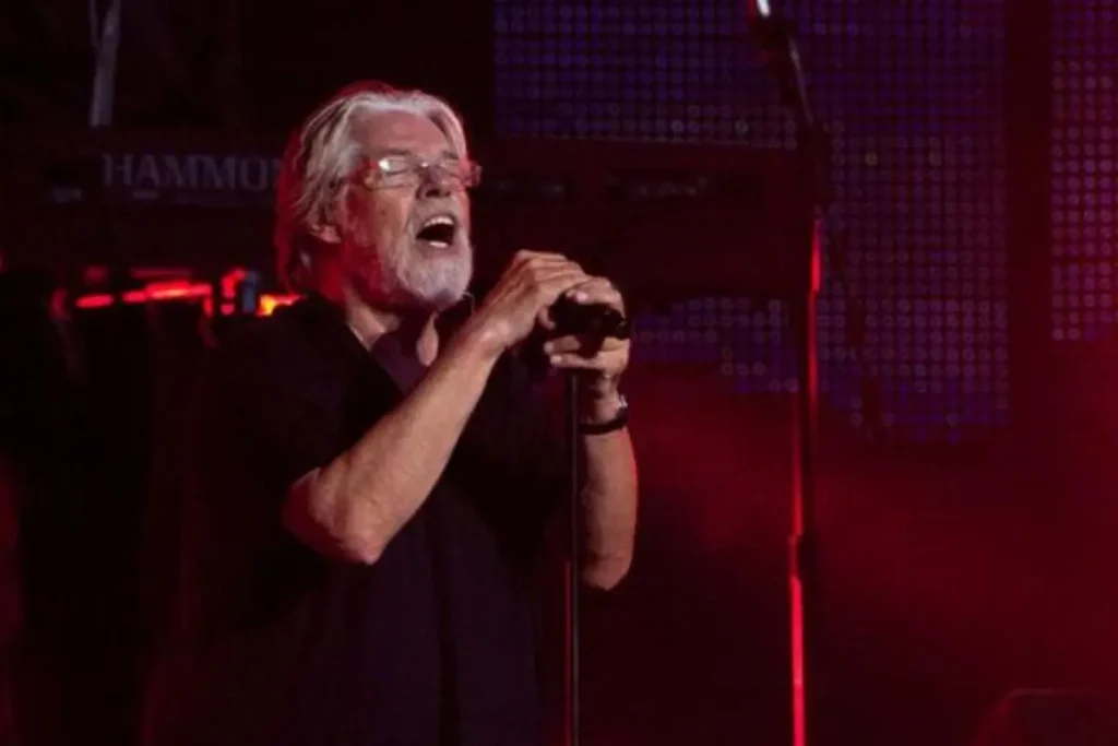 Bob Seger singing passionately into a microphone onstage, wearing glasses and a black shirt under red stage lighting.
