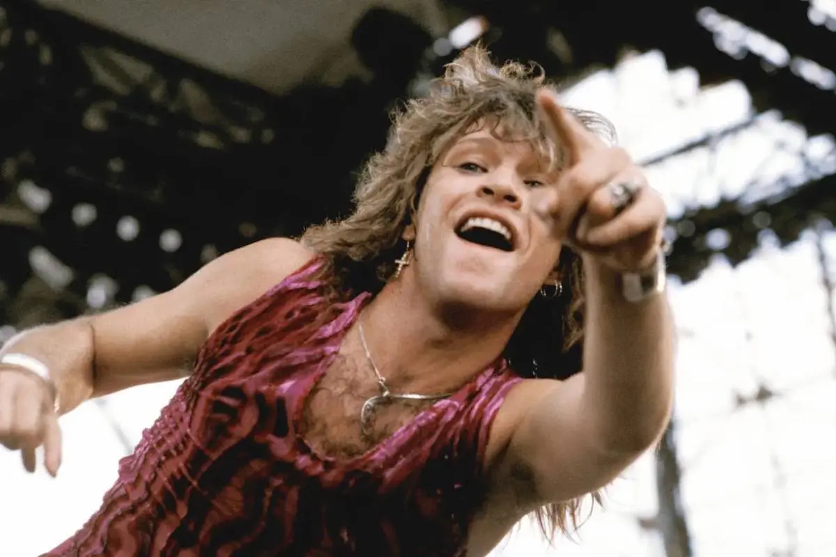 Bon Jovi with long, curly hair leans toward the crowd on an outdoor stage.