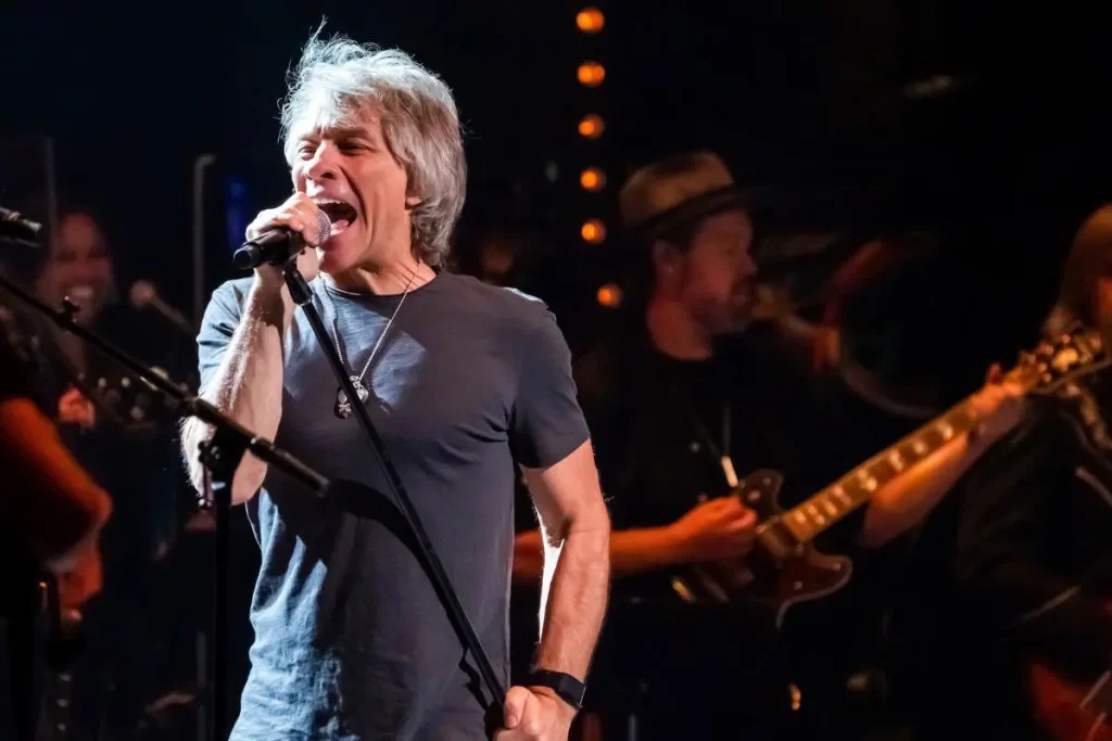 Bon Jovi with gray hair sings passionately into a microphone on a dimly lit stage.