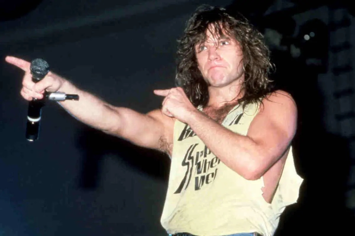 Bon Jovi with long hair and muscular arms points outward while holding a microphone.