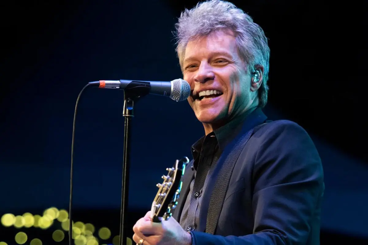 Bon Jovi singing into a microphone and holding a guitar, illuminated by stage lighting during a concert.