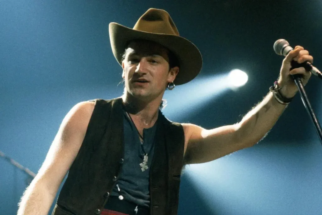 Bono U2 wearing a wide-brimmed hat and sleeveless vest, holding a microphone onstage under bright concert lights.