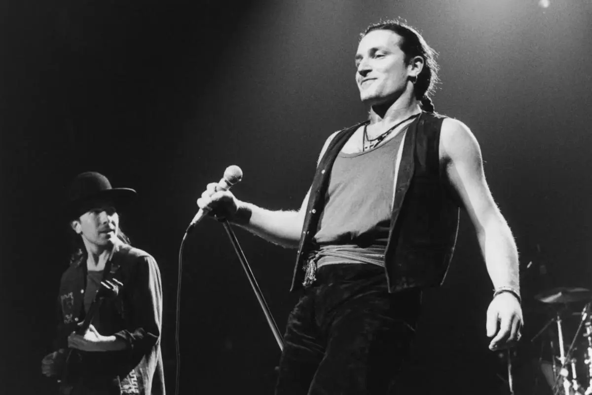 Bono U2 at center stage holding a microphone, with a guitarist visible in the background during a live performance.