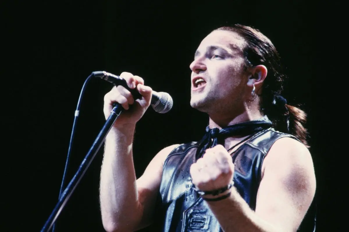Bono U2 with long hair tied back, wearing a sleeveless leather vest, singing passionately into a microphone during a concert.