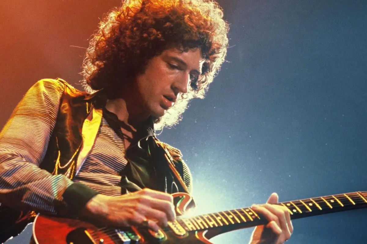 Brian May focused on playing an electric guitar while wearing a striped shirt and dark vest.