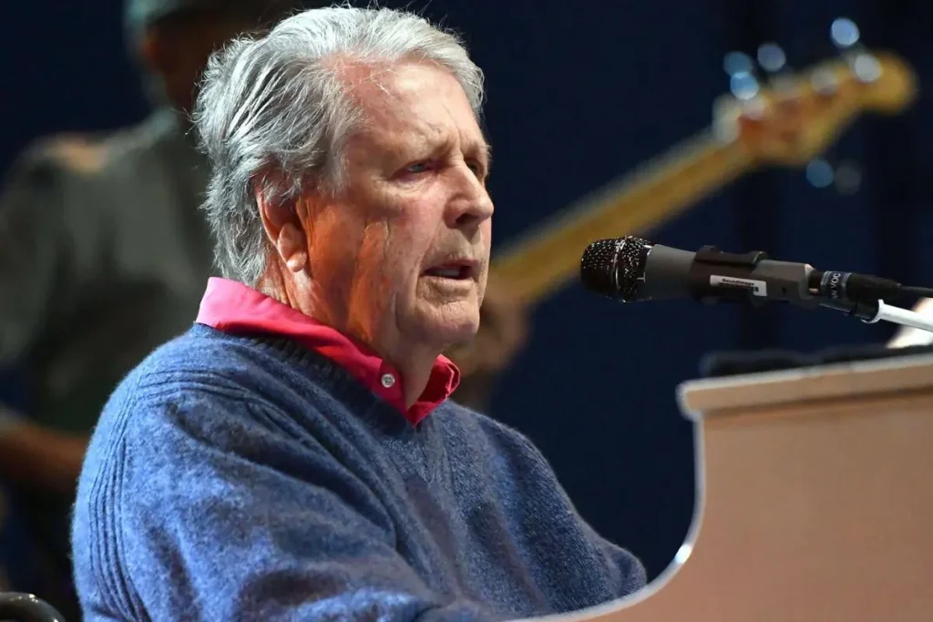 Brian Wilson gray hair sings into a microphone while seated at a white piano onstage.