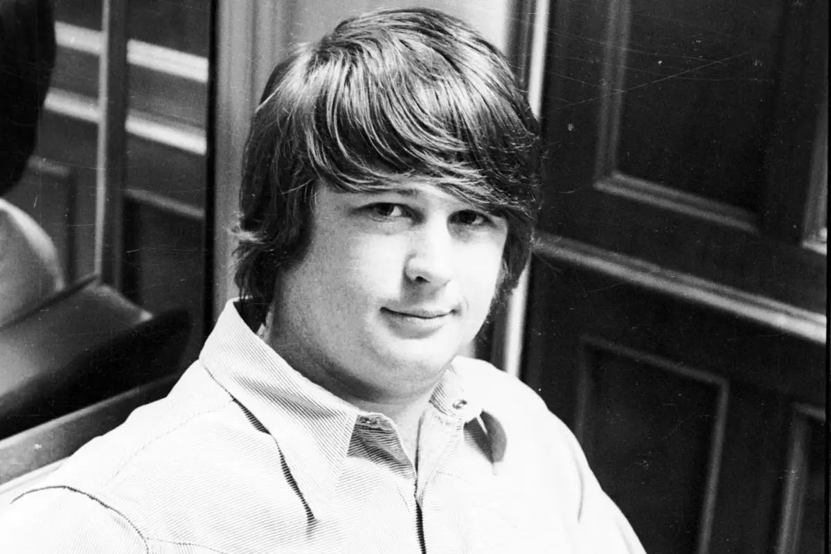 Brian Wilson with medium-length hair seated indoors, looking toward the camera with a calm expression, framed by a doorway and wall panels.