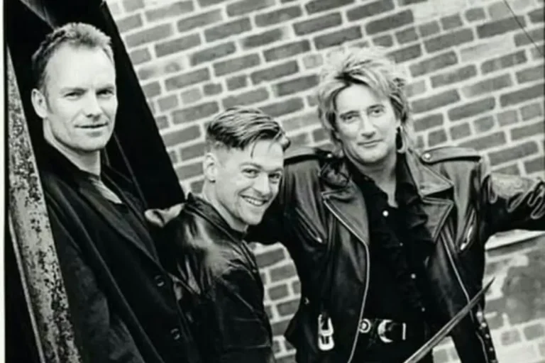 three rock musicians standing against a brick wall, wearing leather jackets and looking toward the camera.
