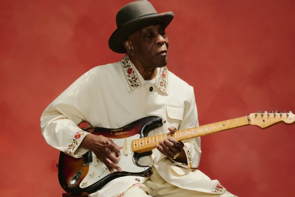 Buddy Guy seated against a red backdrop, playing an electric guitar, highlighting his enduring influence as a blues legend.