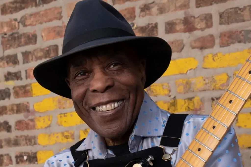 Legendary blues guitarist Buddy Guy smiles while holding an electric guitar, highlighting his enduring influence on blues and rock music.