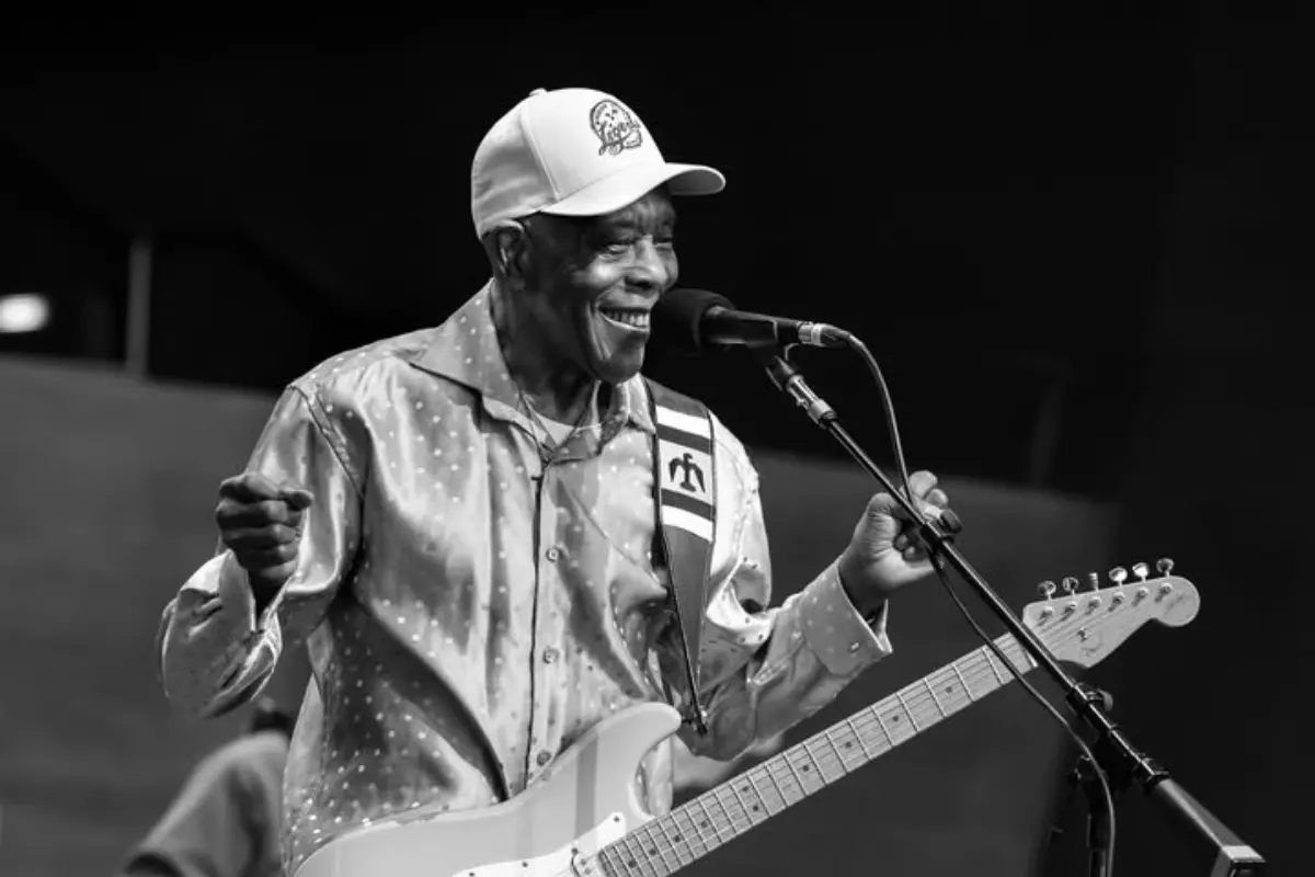 Buddy Guy performs on stage, singing into a microphone while playing electric guitar, capturing the energy of a live blues concert.