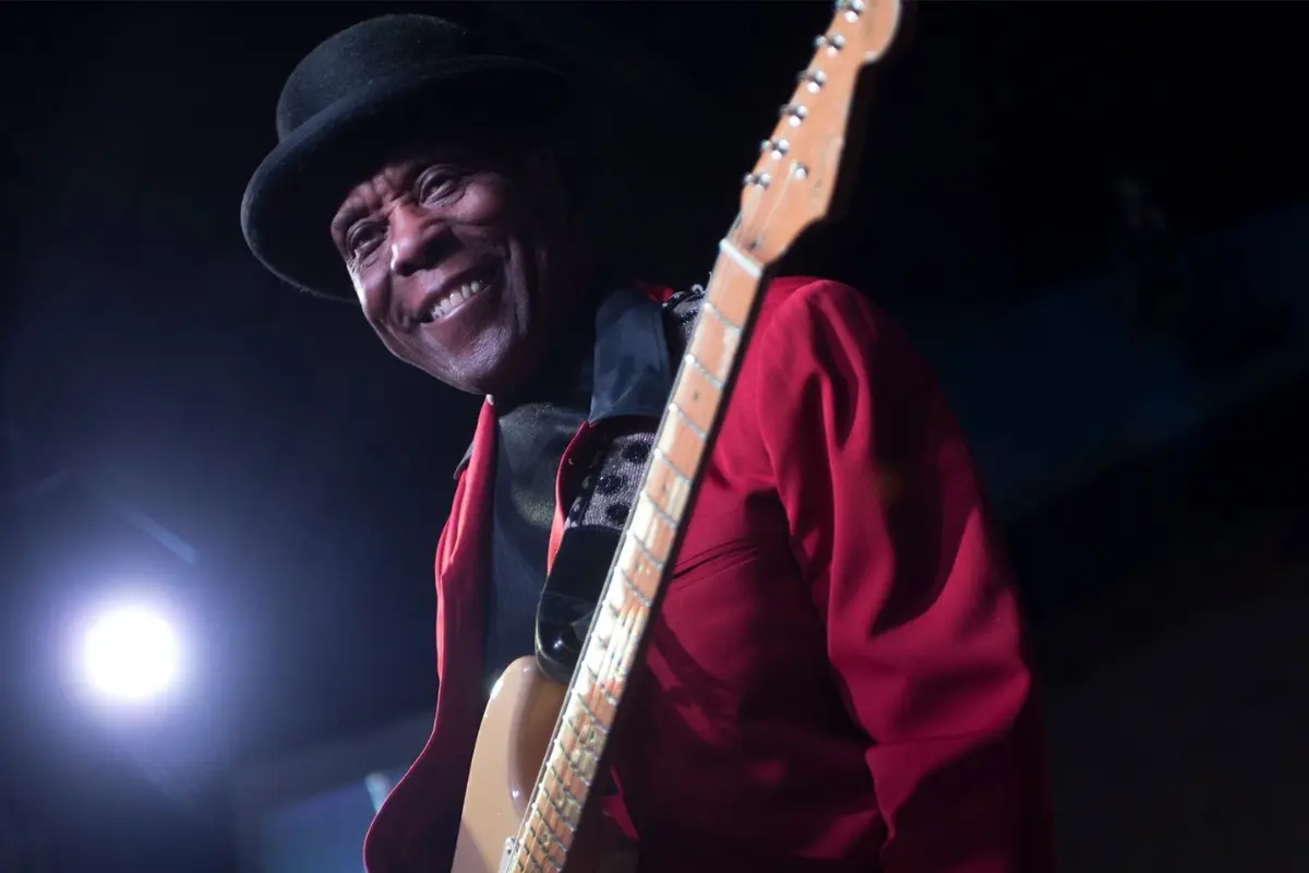 Buddy Guy smiling on stage while playing electric guitar, capturing the joy and energy of a live blues performance.