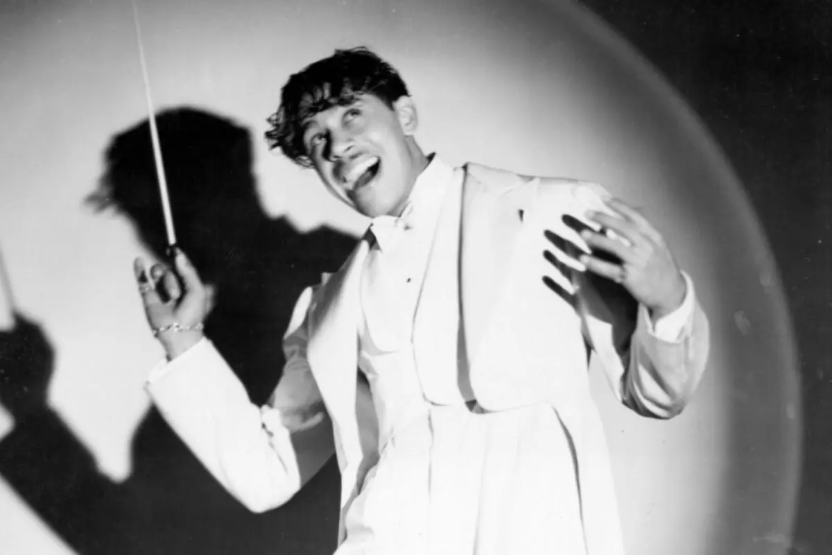 Cab Calloway holding a baton and smiling broadly under a spotlight, casting a dramatic shadow on the backdrop.