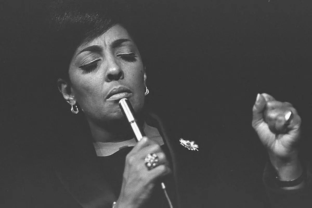 Carmen McRae singing into a handheld microphone, eyes closed, wearing earrings and a dark outfit, conveying deep emotion.