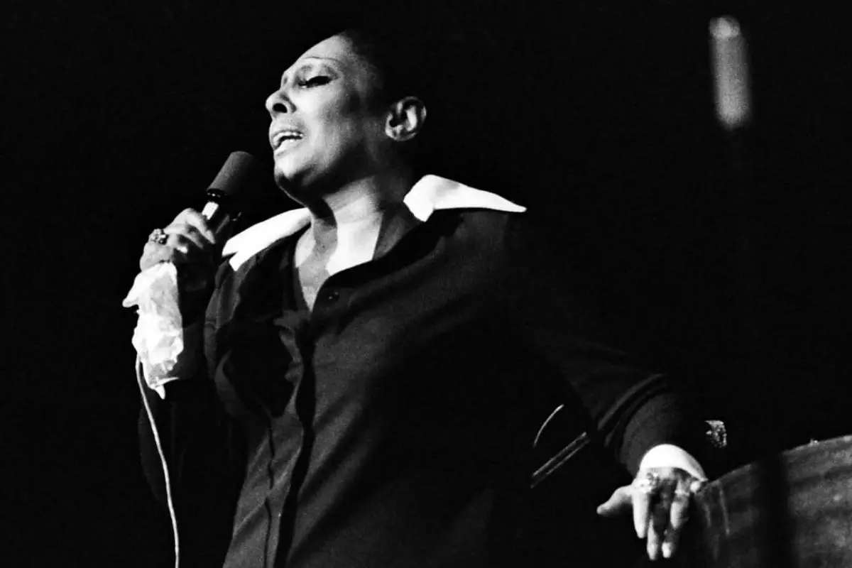 Carmen McRae holding a microphone in one hand while leaning against a piano, captured mid-song with dramatic lighting.