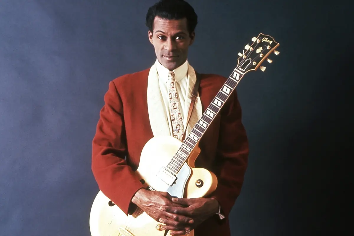 Chuck Berry holding an electric guitar against a dark backdrop, presenting a poised, confident image of a rock and roll pioneer.