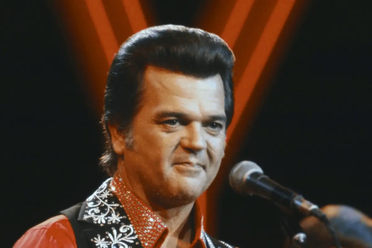 Conway Twitty performing on stage, wearing a red embellished shirt and black vest, framed by warm stage lighting.