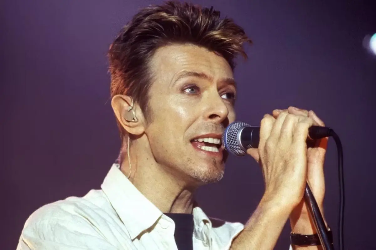 David Bowie singing into a handheld microphone during a live performance, wearing a white shirt.