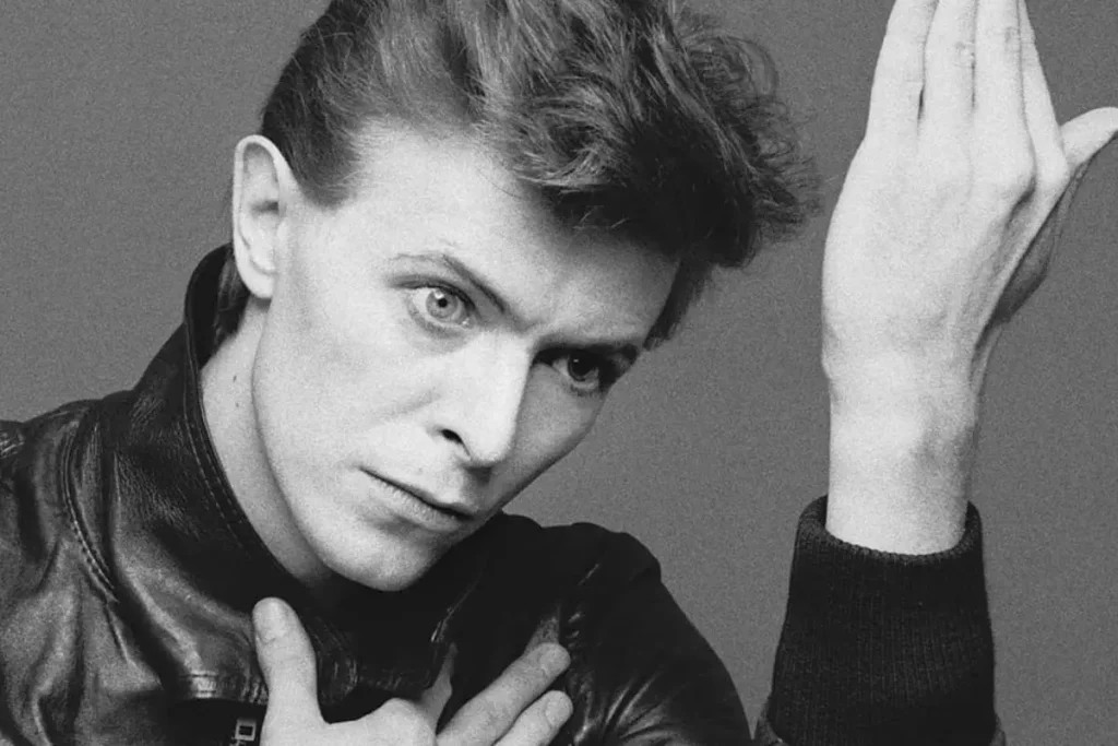 Bowie, Bolan, and 17 Years of Silence: The Glam Rock Friendship Nobody Saw David Bowie with short styled hair, wearing a dark jacket, gazing intensely toward the camera with one arm raised.