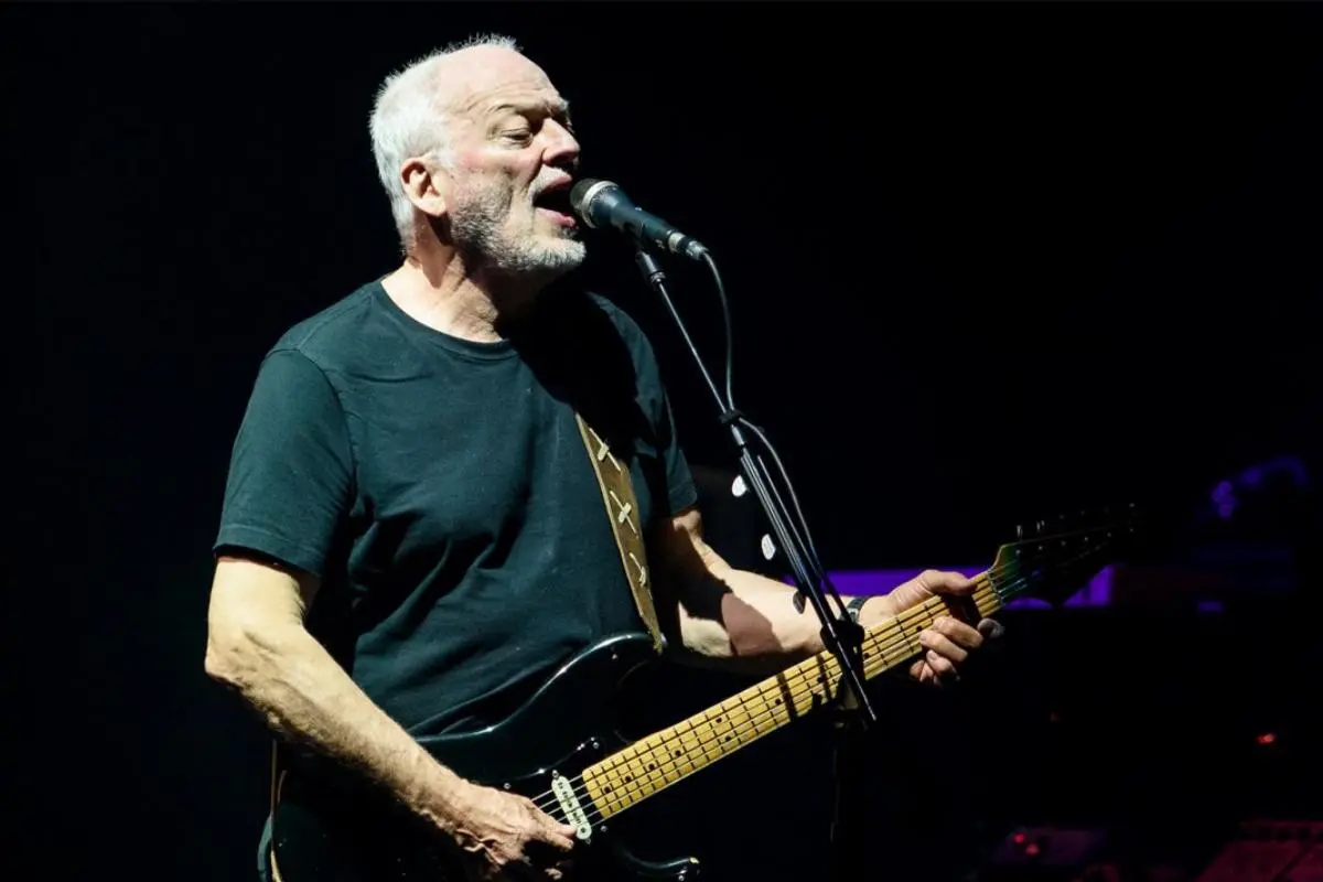 David Gilmour performing live on stage, singing into a microphone while playing electric guitar under dark concert lighting.