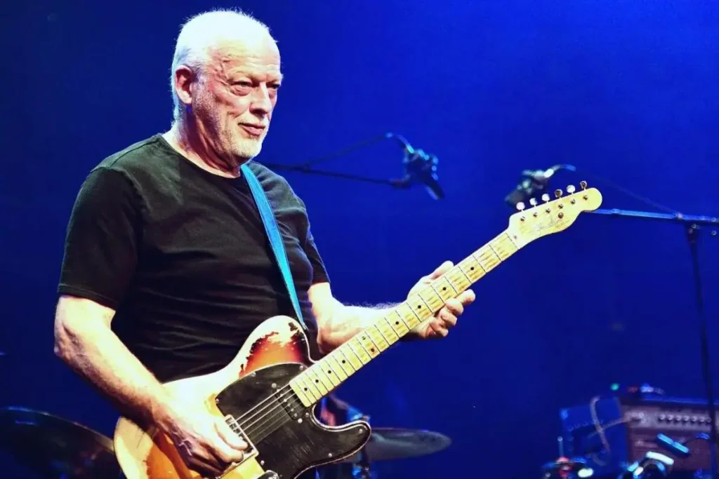 David Gilmour playing electric guitar on stage, wearing a black T-shirt and standing under blue stage lights during a live performance.