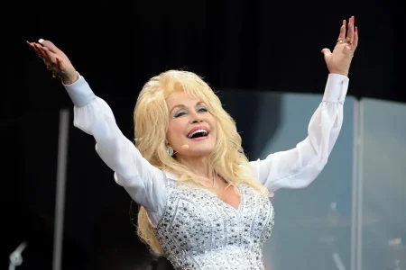Dolly Parton performing on stage with arms raised, wearing a white embellished outfit and smiling toward the audience.
