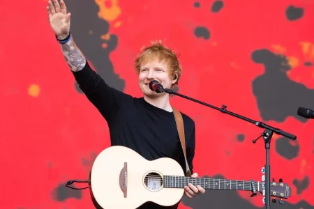 Ed Sheeran waves to the crowd while holding an acoustic guitar on stage, standing in front of a bright red backdrop during a live performance.