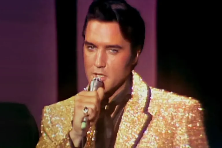 Elvis Presley wearing a gold sequined jacket, singing into a handheld microphone on stage under warm lighting.