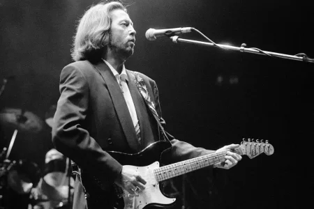 Eric Clapton singing into a microphone while playing electric guitar during a live performance.
