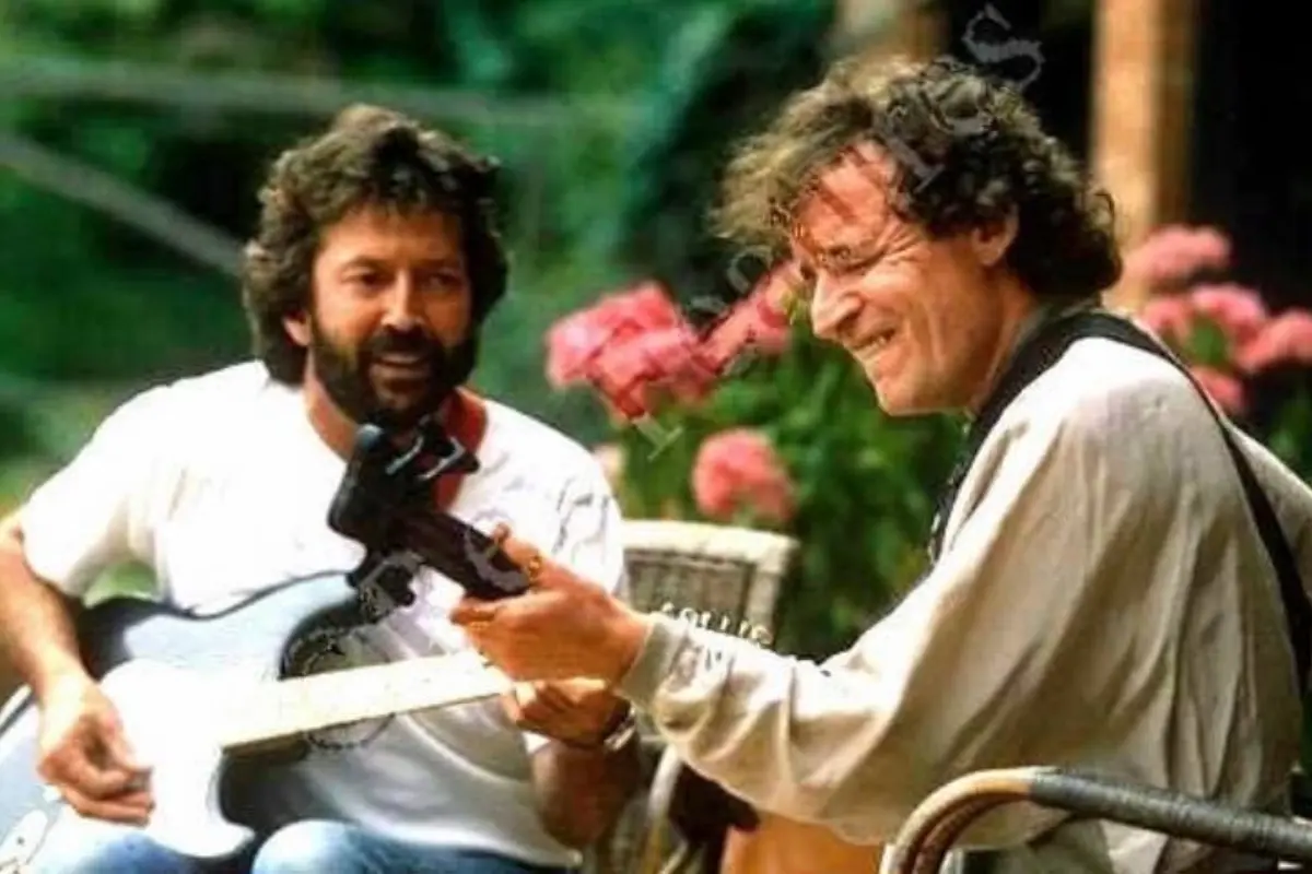 Eric Clapton and Jack Bruce sit outdoors, smiling and playing electric guitars together in a relaxed, informal setting.