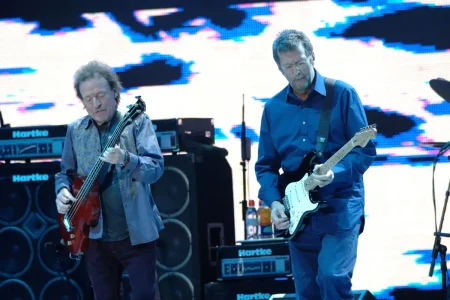 Eric Clapton and Jack Bruce play electric guitars side by side during a large live concert, with amplifiers and a bright stage backdrop behind them.