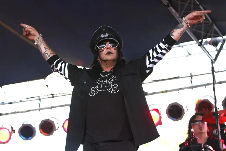 Faster Pussycat wearing black clothing with striped sleeves and a military-style hat under concert lighting.