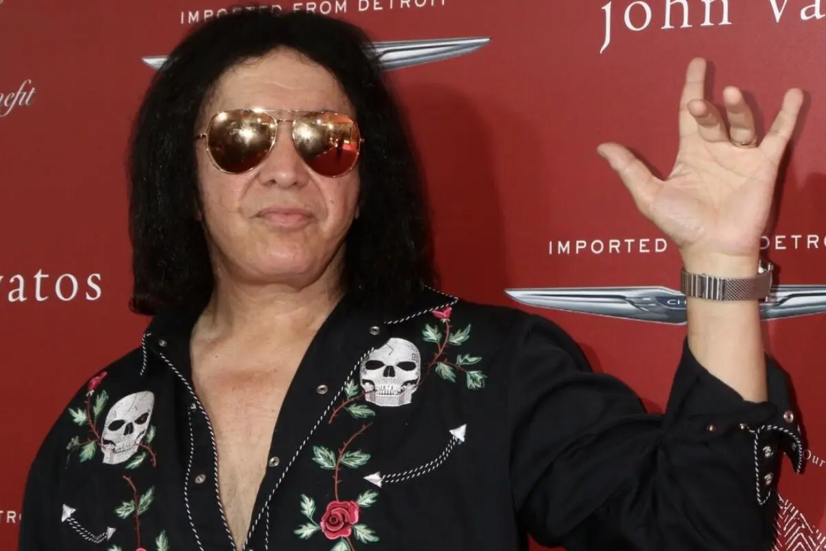 Gene Simmons wearing sunglasses and a black embroidered shirt with skull designs, raising his hand on a red carpet backdrop.