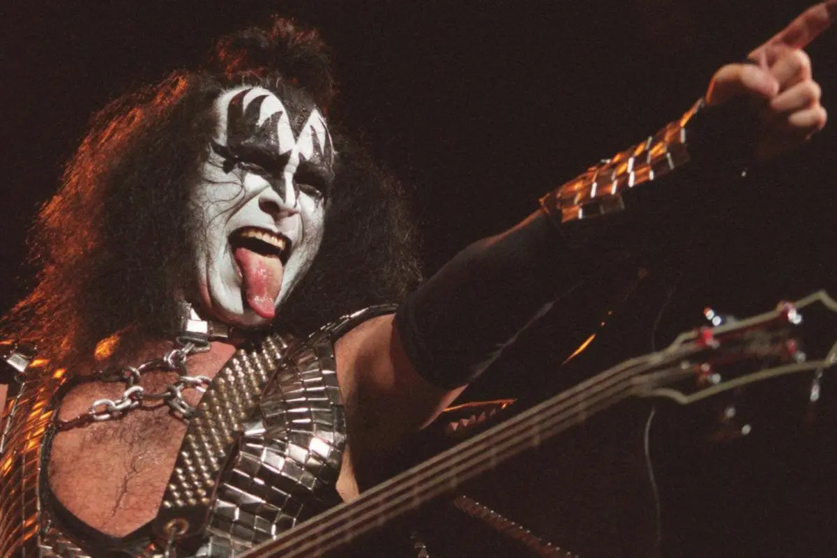 Gene Simmons performing live on stage in full black-and-white Demon makeup, pointing toward the crowd while playing bass guitar.