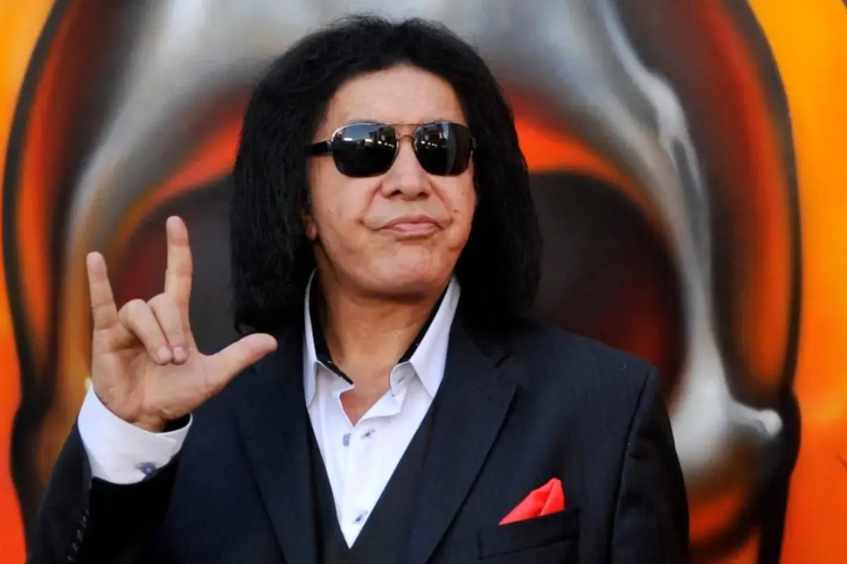Gene Simmons in a black suit and sunglasses making the ‘rock on’ hand gesture at a public event.