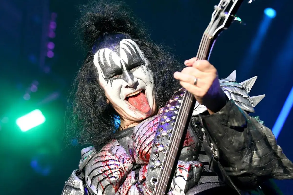 Gene Simmons playing bass guitar on stage with iconic Demon face paint and extended tongue under concert lighting.