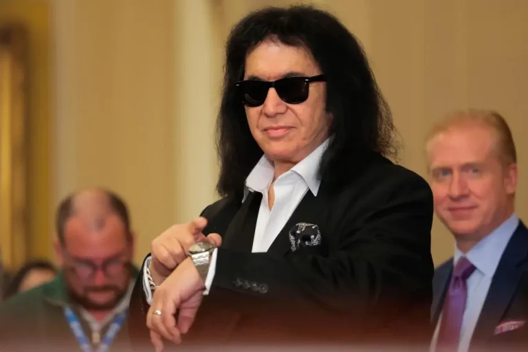 Gene Simmons dressed in a black suit and white shirt, wearing sunglasses and checking his watch while standing indoors.