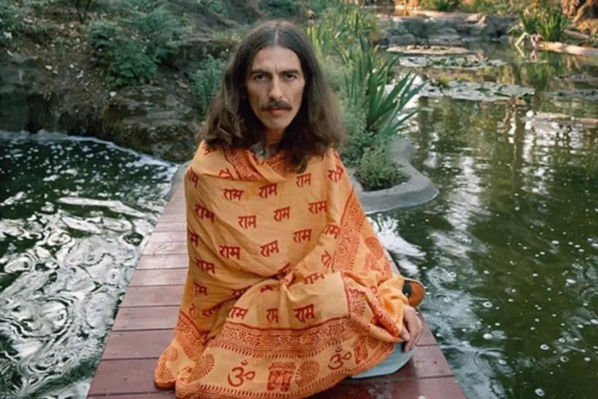 George Harrison seated outdoors, wrapped in a saffron robe beside a pond.