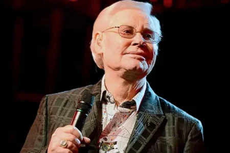 George Jones holds a microphone under stage lighting, wearing glasses and a patterned jacket.