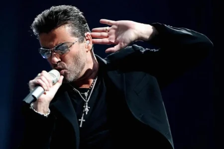 George Michael holding a microphone close to his mouth with one hand raised to his ear.