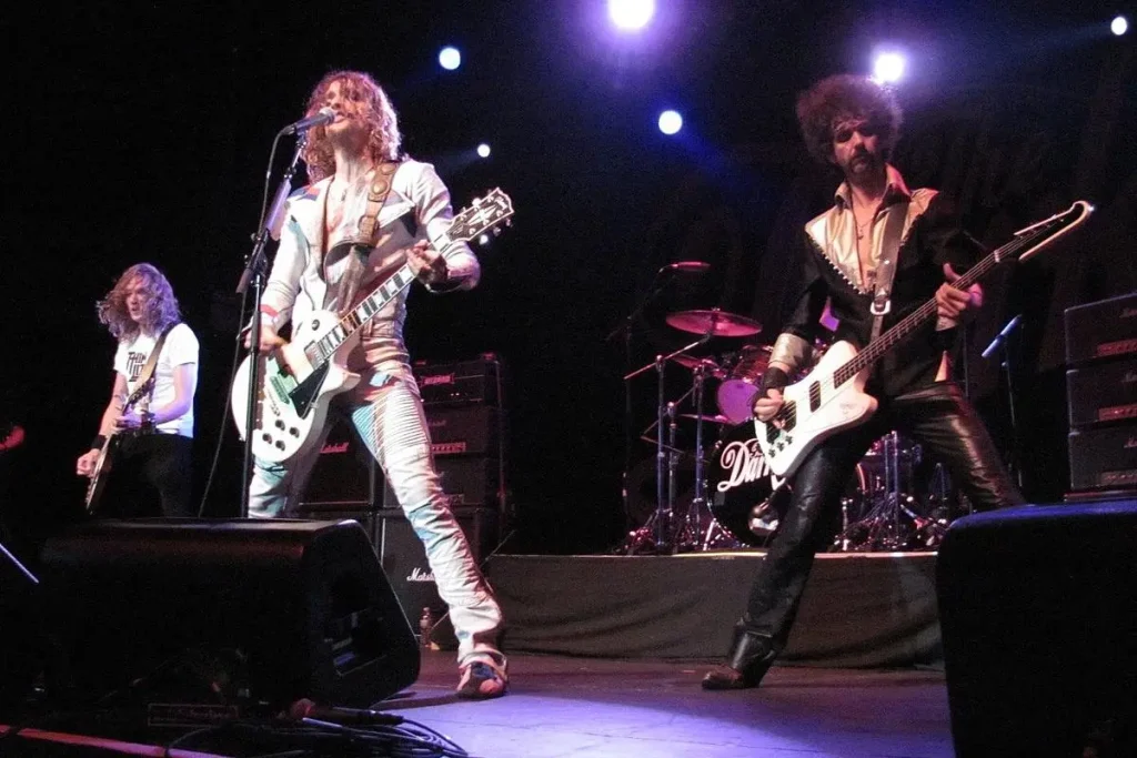 Glam Rock performing live on stage under bright concert lights, with multiple guitarists and a bassist playing electric guitars in front of a large audience.