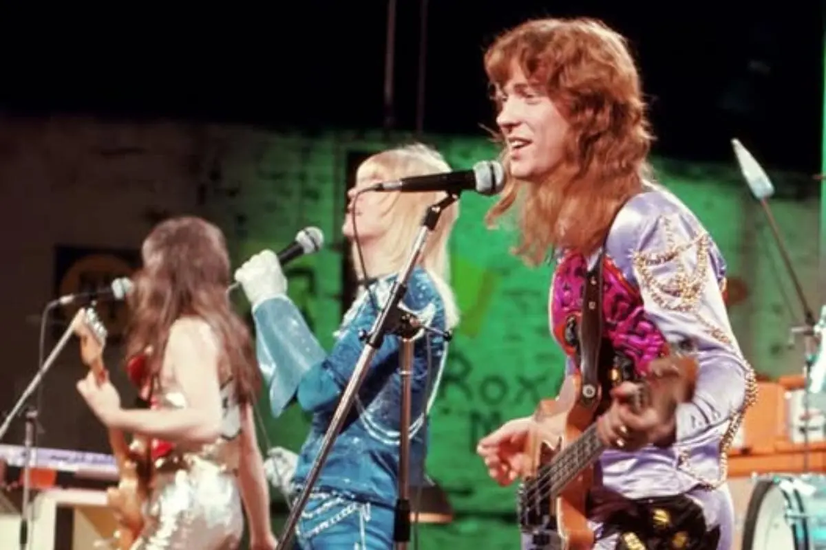 Glam Rock performing on stage, wearing colorful, shiny outfits while singing into microphones and playing electric guitars.