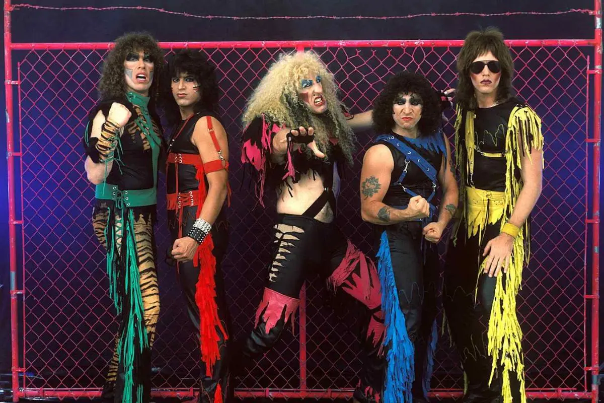Glam Rock posing in dramatic stage costumes with bold makeup, ripped leather outfits, and bright colors, standing in front of a chain-link fence backdrop.