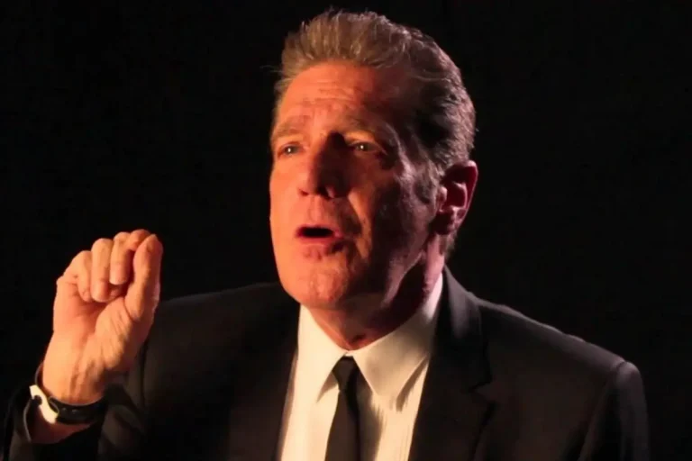 Glenn Frey in a dark suit and tie speaks emphatically against a black background.