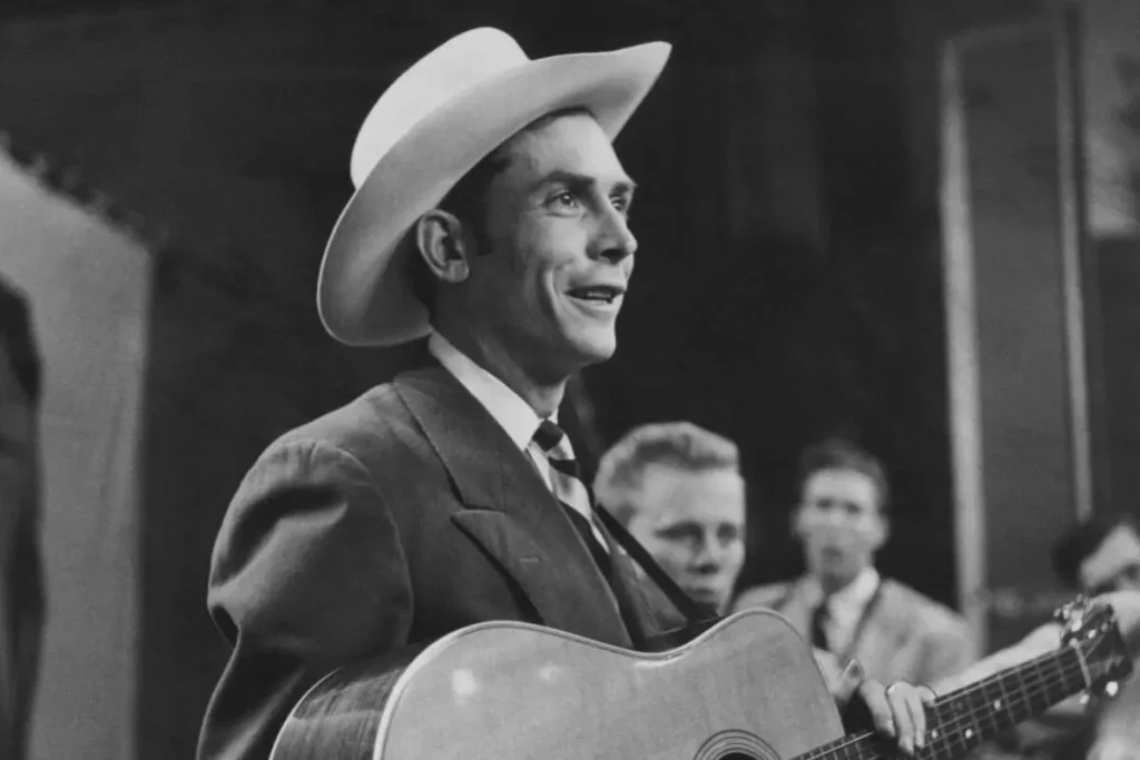 Hank Williams playing an acoustic guitar onstage near a vintage microphone.
