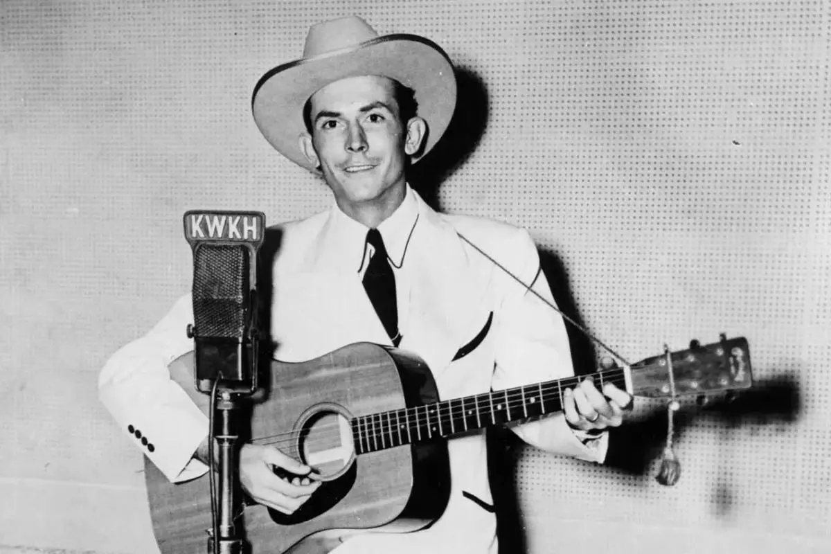 Hank Williams standing at a microphone labeled “KWKH” while playing an acoustic guitar.