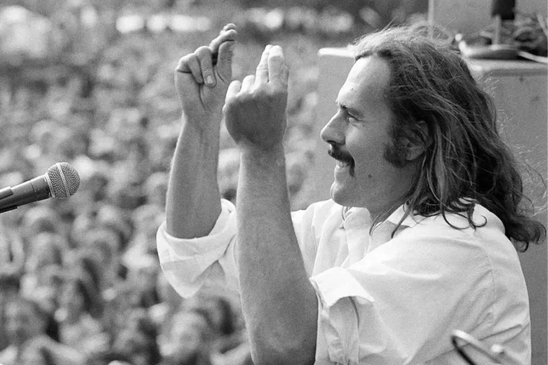 Hot Rod Lincoln in profile on stage facing a large outdoor crowd; he smiles and holds both hands up with fingers pinched together.