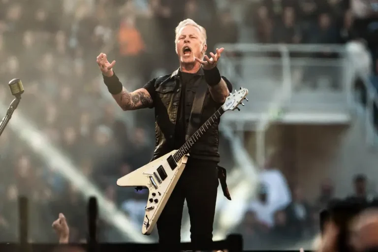 James Hetfield performs on a large stage, arms raised toward the crowd with guitar strapped across his body.