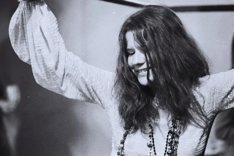 Janis Joplin performing live, smiling and singing passionately into a handheld microphone under stage lighting.