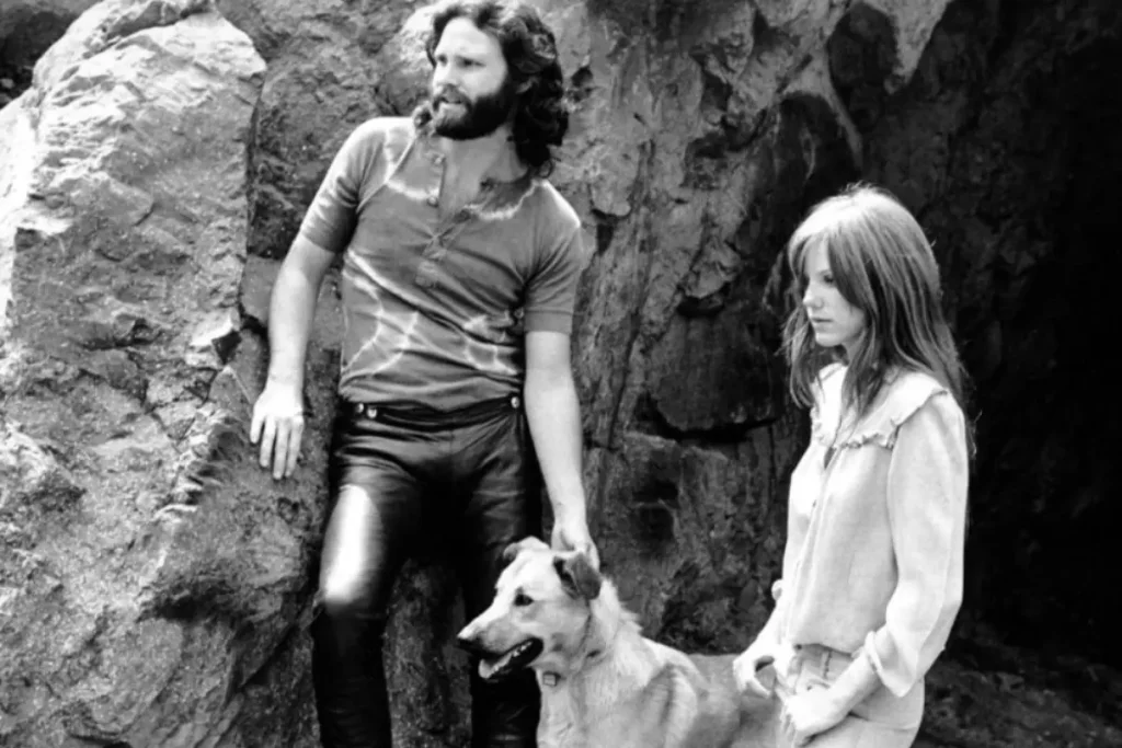 Jim Morrison stands beside Pamela Courson with their dog near a rocky outdoor setting, capturing a candid glimpse of their shared life away from the stage.